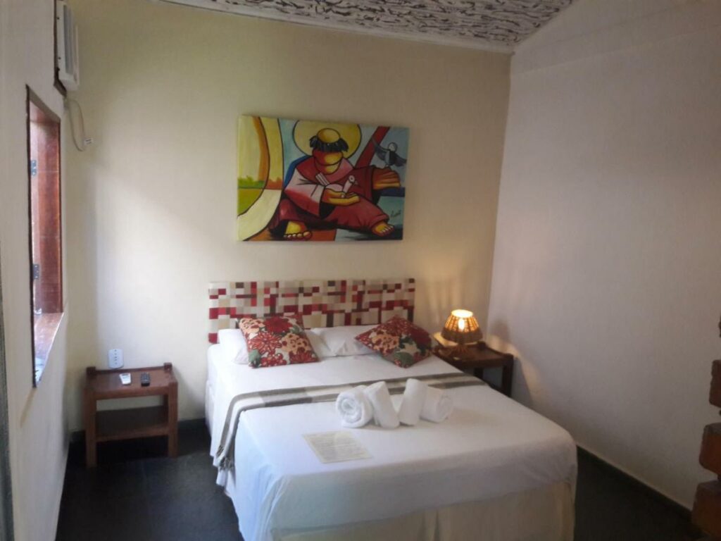 Quarto da Pousada Macunaíma. A cama de casal tem lençol, travesseiros e toalhas brancas em cima da cama. Duas almofadas coloridas estão contra os travesseiros, e um quadro colorido na parede acima.
Há mesinhas de cabeceira dos dois lados da cama, estando um abajur aceso do lado direito e dois controles do lado esquerdo. Há uma janela na parede esquerda e um ar-condicionado ao lado.