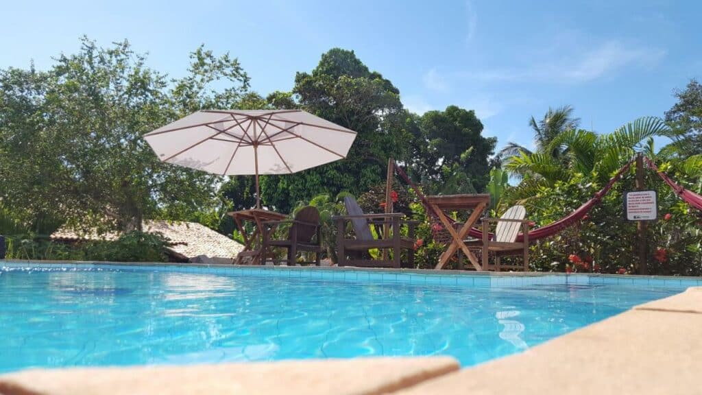 Piscina da Pousada Tubarão, uma das recomendações de pousadas em Arraial D'Ajuda. Há cadeiras e mesas de madeira na beira da piscina, e um guarda-sol aberto. Algumas árvores podem ser vistas ao fundo.