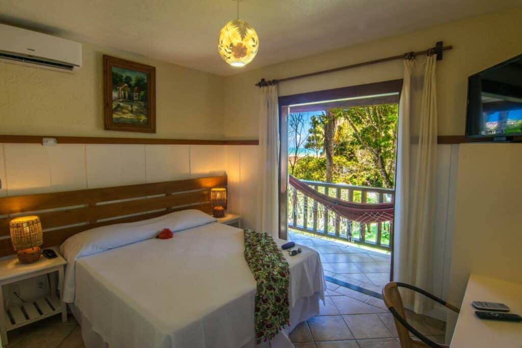 Quarto da Manacá Pousada Parque. Há uma cama com lençol e travesseiros brancos com uma canga florida em cima. Há abajures nas mesinhas de cabeceira dos dois lados da cama. Um ar-condicionado está ao lado de um quadro na parede acima da cama.
Há uma mesa de trabalho com cadeira em frente, e uma televisão na parede lateral.
Na varanda com vista para árvores há uma rede.