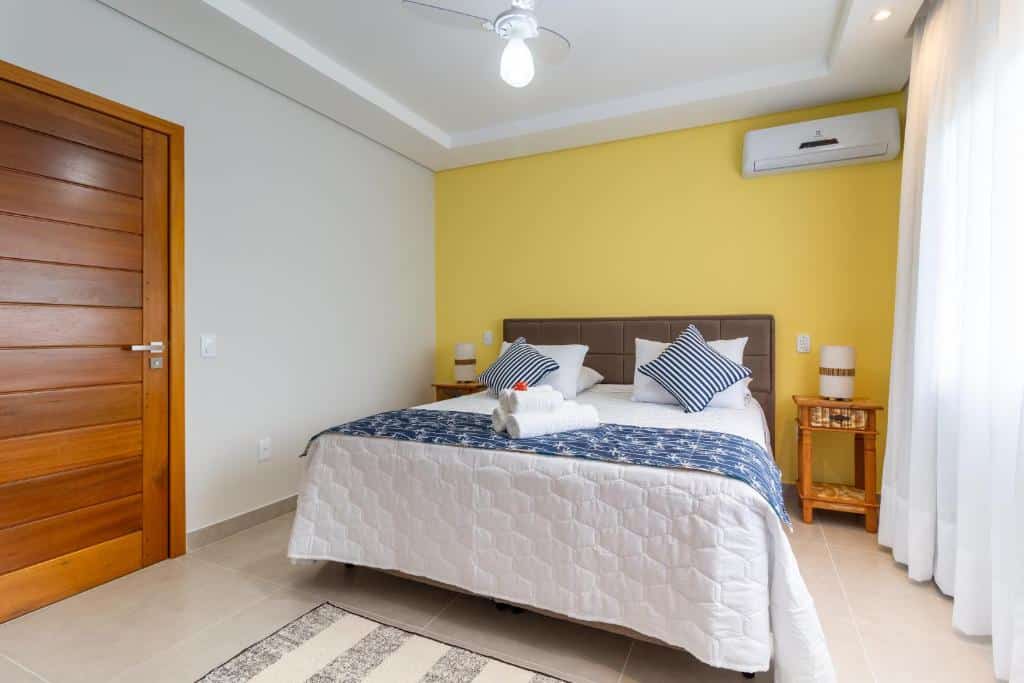 Quarto da Pousada Casa Cajá, uma das recomendações de pousadas em Arraial D'Ajuda. A cama de casal tem lençol e travasseiros azul e brancos, com algumas toalhas dobradas em cima. Há abajures nas mesinhas de cabeceira dos dois lados da cama, e um tapete em frente a ela. A janela está na parede direita com cortinas brancas que vão até o chão, a porta marrom está na parede esquerda e há um aparelho de ar-condicionado na parede acima da cama.