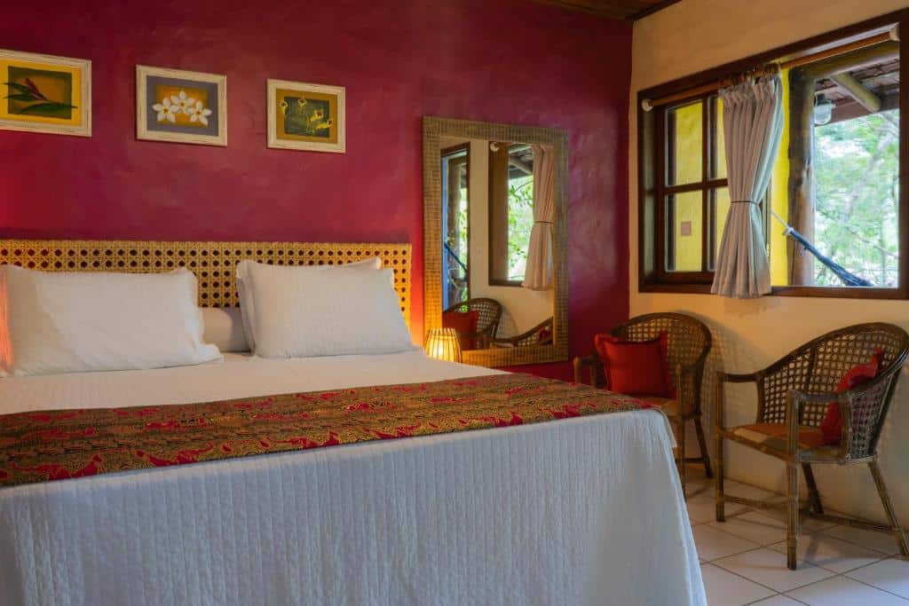Quarto da Atmosphera Pousada. A cama tem lençol e travesseiros brancos e colcha rosa. Há três quadrinhos na parede acima, e um espelho do lado direito da cama. É possível ver um abajur ao lado da cama.
Uma janela do lado direito do quarto tem vista para o jardim e uma rede pendurada na varanda. Há duas cadeiras com almofadas embaixo da janela.