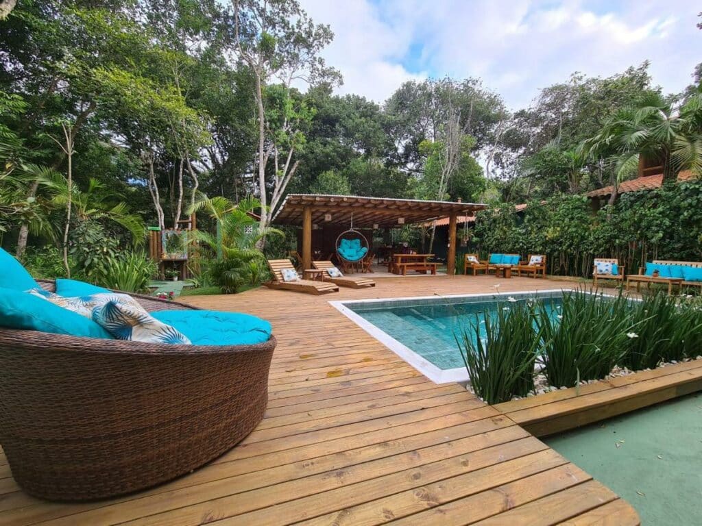 Área da piscina da Quintessência Pousada de Charme, uma das recomendações de pousadas em Arraial D'Ajuda. Há várias árvores ao redor, e espreguiçadeiras e assentos com almofadas azuis estão espalhadas pela área. Ao fundo há uma área coberta.