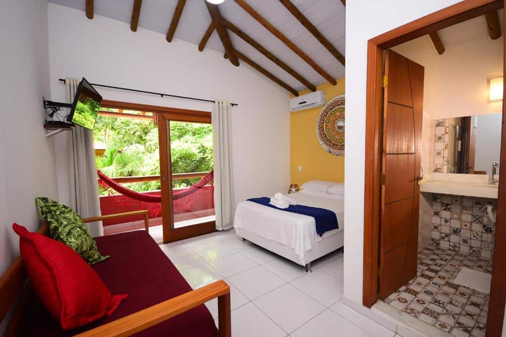 Quarto da Quintessência Pousada de Charme. A cama tem lençol e travesseiros brancos com algumas toalhas brancas dobradas em cima de um cobertor azul. Há abajures nas mesinhas de cabeceira dos dois lados, uma mandala na parede acima e um ar-condicionado ao seu lado.
Uma televisão encara a cama do outro lado, com uma porta que leva à varanda com rede ao lado.
Há um sofá com almofadas encarando a porta do banheiro.