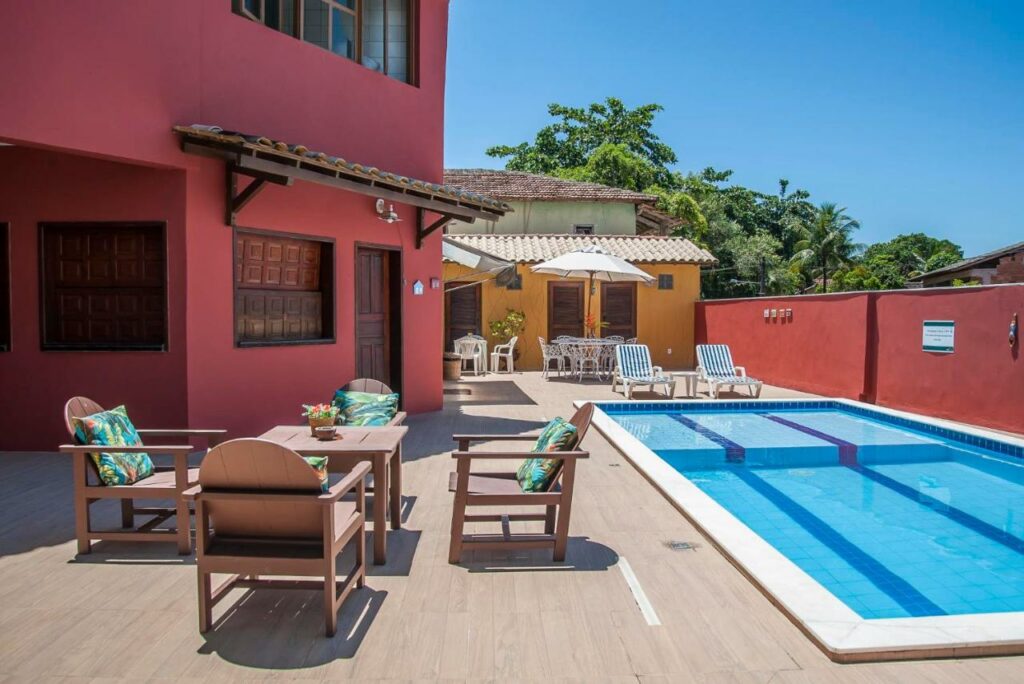 Área da piscina da Pousada Manhã Dourada, uma das recomendações de pousadas em Arraial D'Ajuda. Há mesinhas com cadeiras de madeira e guarda-sol ao redor, e duas espreguiçadeiras encaram a piscina.