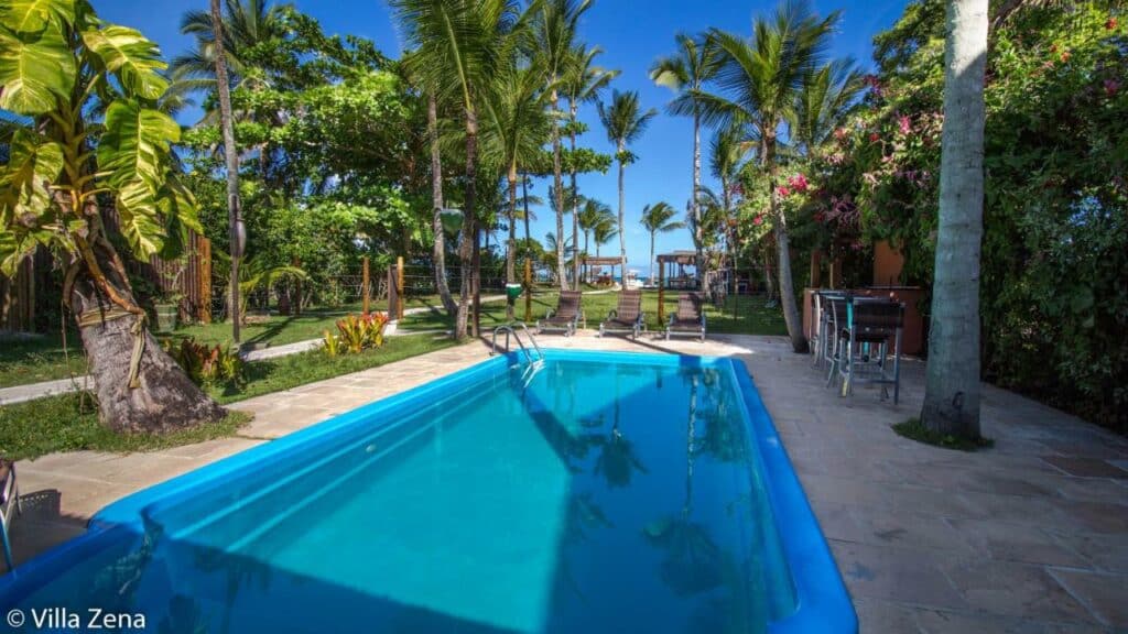Piscina da Pousada Villa Zena, uma das recomendações de pousadas em Arraial D'Ajuda. Há diversas árvores ao redor, e algumas espreguiçadeiras e mesinhas com cadeiras ao longo da extensão da piscina. Ao fundo é possível ver a praia.