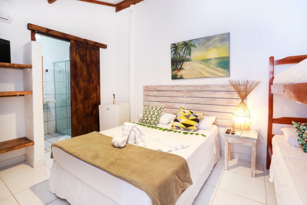 Quarto da Pousada do Mel, uma das recomendações de pousadas em Arraial D'Ajuda. A cama tem lençol e travesseiros coloridos, e algumas toalhas brancas dobradas em cima. Há um quadro acima da cabeceira de madeira, e do lado direito uma mesinha de cabeceira com abajur. Ao lado há uma beliche.
Do outro lado da cama há um frigobar, e a porta do banheiro está ao lado. Há algumas prateleiras vazias do outro lado da porta.
