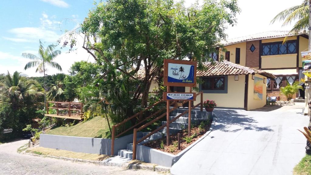 Fachada da Pousada Vento Sul, uma das recomendações de pousadas em Arraial D'Ajuda. Há algumas árvores ao redor, e uma escadaria que leva à pousada amarela. A placa que indica o nome do hotel está ao lado da escada.