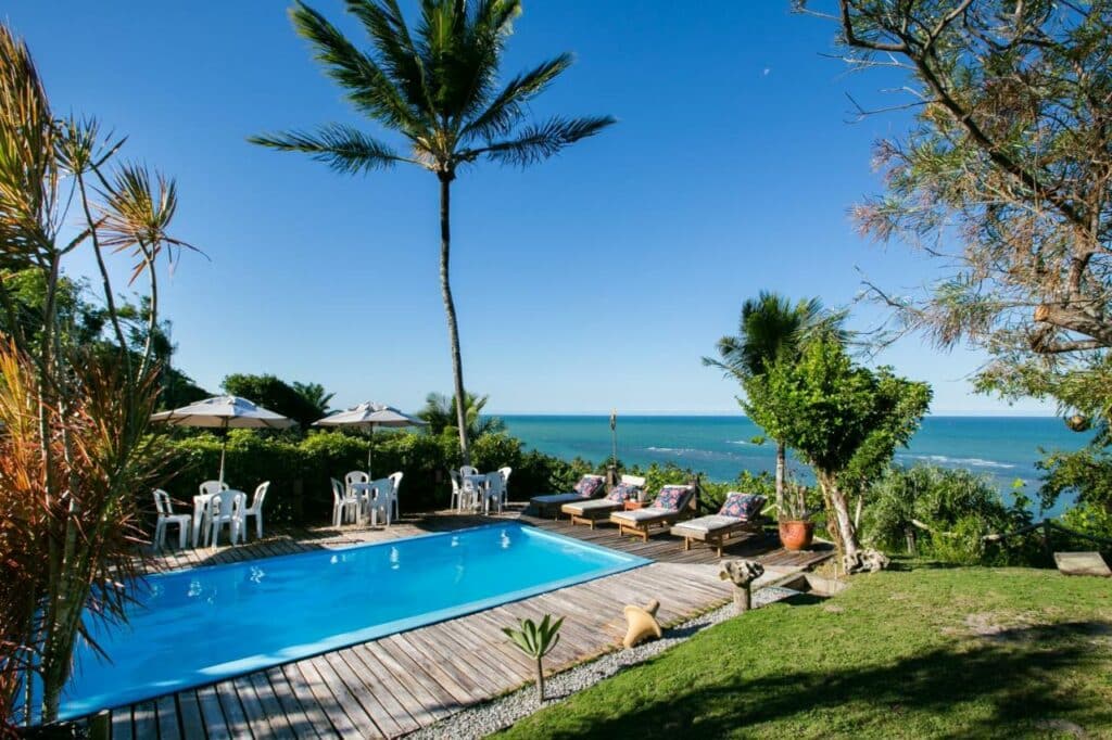 Piscina da Pousada Jakuara, uma das recomendações de pousadas em Arraial D'Ajuda. Há árvores e grama ao redor, e algumas mesas com cadeiras e espreguiçadeiras ao redor da piscina. É possível ver o mar ao fundo.