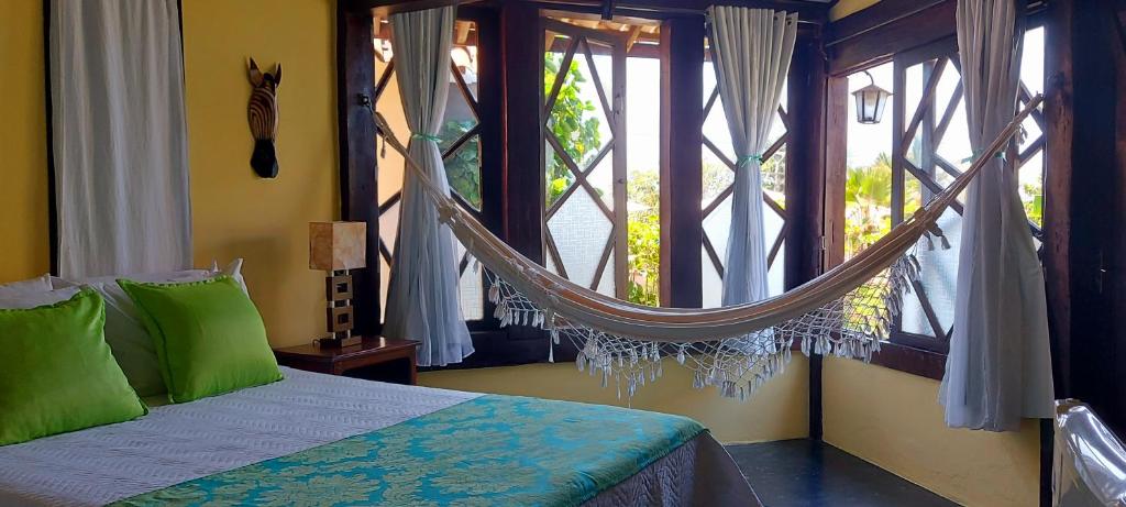 Quarto da Pousada Vento Sul. A cama de casal tem lençol e travesseiros verde e brancos. Ao lado da cama há uma mesinha de cabeceira com abajur, e uma decoração de parede em formato de cabeça de zebra.
A parede ao lado é composta por janelas, e uma rede está pendurada de ponta a ponta.