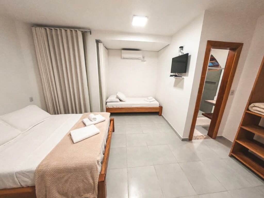 Quarto da Amarilis Pousada Arraial d'Ajuda, uma das recomendações de pousadas em Arraial D'Ajuda. Uma cama de casal com lençol, toalhas e travesseiros brancos está encostada na parede esquerda. Há uma cama de solteiro ao lado com um ar-condicionado acima. Uma televisão está na parede de frente.
Na lateral direita é possível ver um ármario e uma porta que leva ao banheiro. Há duas janelas com cortinas no quarto.
