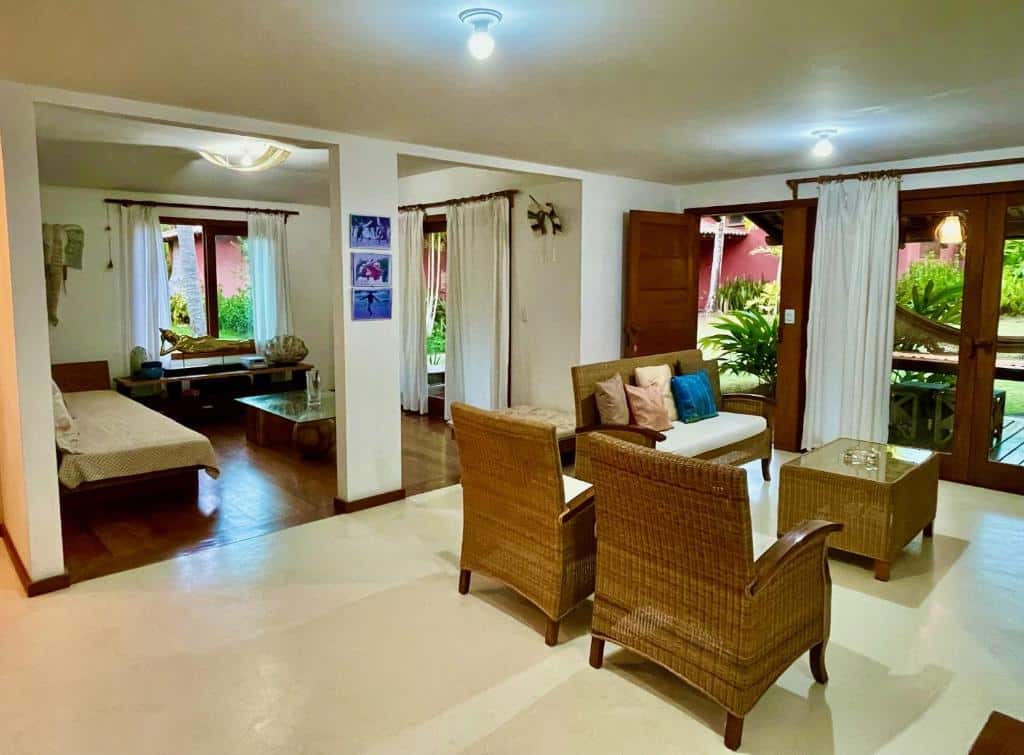 Sala de estar de um apartamento da Charm Bahia Residencias. Há sofás e poltronas com uma mesinha de centro na sala, e uma porta de vidro tem vista para a rede na varanda.
Ao fundo da imagem há uma entrada que leva a um espaço com sofá-cama e uma mesa de vidro.