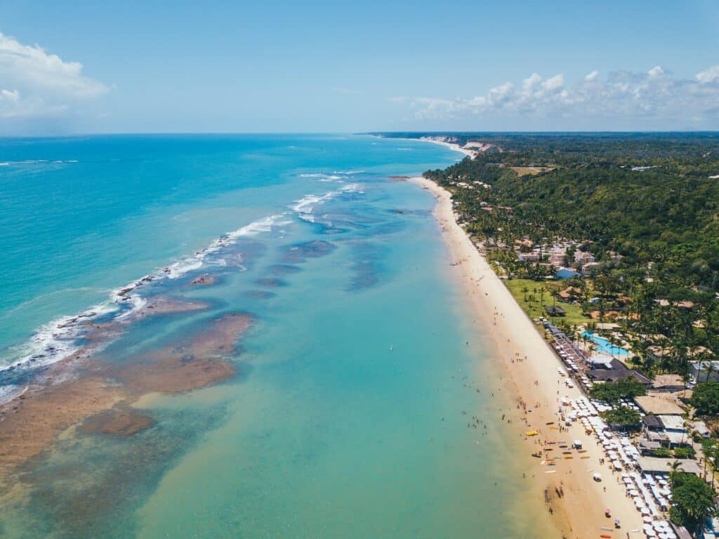 Vista aérea da Praia do Mucugê. Do lado esquerdo está o mar, e do direito a costa com várias casas e árvores. Algumas pessoas estão ao longo da faixa de areia. - Foto: J. Balla Photography via Unsplash
