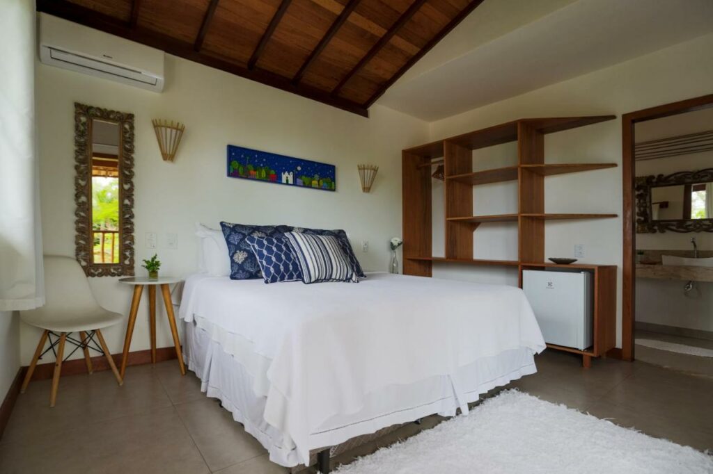 Quarto da Pousada Jakuara. A cama tem lençol branco e travesseiros azuis. Há uma luminária na parede acima da mesinha de cabeceira dos dois lados, e um quadrinho no centro.
Uma cadeira branca está do lado esquerdo da foto com um espelho na parede atrás e um ar-condicionado acima.
Na parede direita há uma estante com divisórias e um frigobar, além de uma porta que leva ao banheiro.