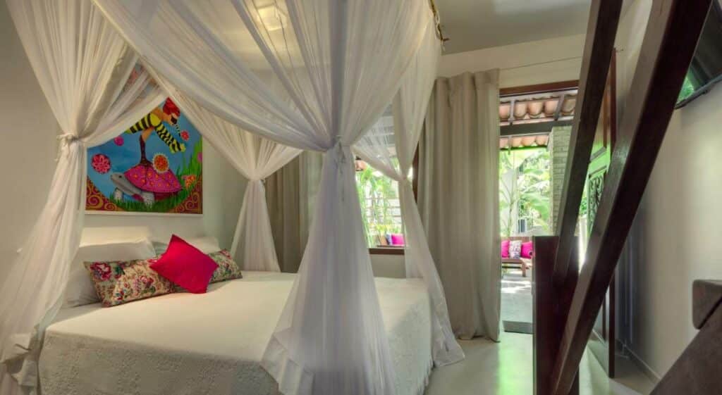 Quarto da Pousada Água de Coco. Uma cama branca com travesseiros rosa tem um mosquiteiro branco preso ao redor. Há um quadro colorido na parede acima da cama, e uma escadaria na sua frente.
Ao fundo é possível ver uma janela e uma porta que leva à varanda com um sofá com almofadas cor de rosa.