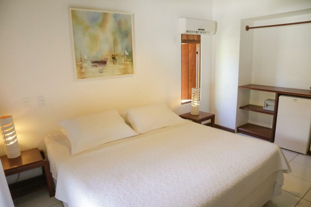 Quarto da Pousada Coisa e Tao. A cama com lençol e travesseiros brancos tem mesinhas de cabeceira dos dois lados com um abajur em cima. Há um quadro na parede acima, e um ar-condicionado acima de um espelho está na parede atrás da mesinha do lado direito.
Há uma estante com prateleiras e um frigobar no canto direito do quarto.