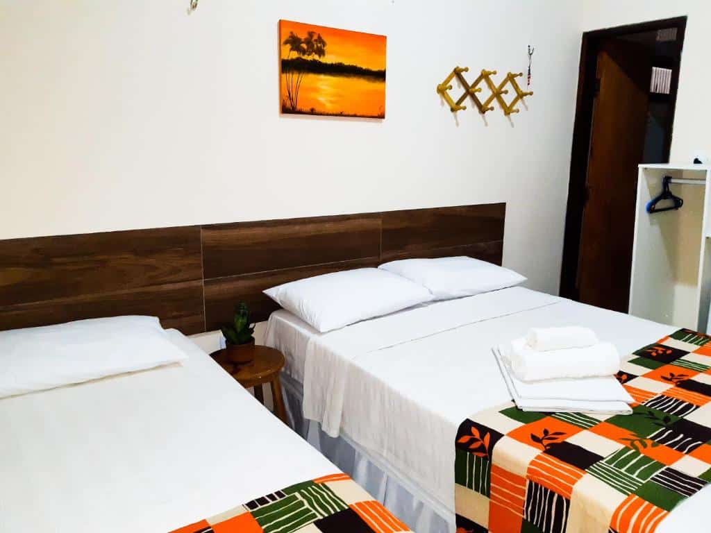 Duas camas bem arrumadas com lençóis brancos, cobertores coloridos e uma toalha dobrada em um quarto simples e bem iluminado, na Pousada do Porto, para representar pousadas em Barreirinhas.