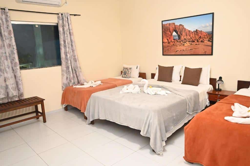 Um quarto na Pousada Paraíso dos Lençóis com três camas, uma janela com cortinas, um banco e uma foto de paisagem emoldurada na parede, para representar pousadas em Barreirinhas.