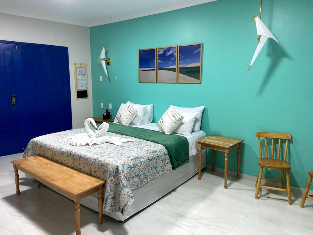 Uma cama bem arrumada com detalhes em verde, móveis de madeira, parede azul-petróleo e decorações de guindaste de origami em um quarto bem iluminado, na Pousada Vila de Charme, para representar pousadas em Barreirinhas.
