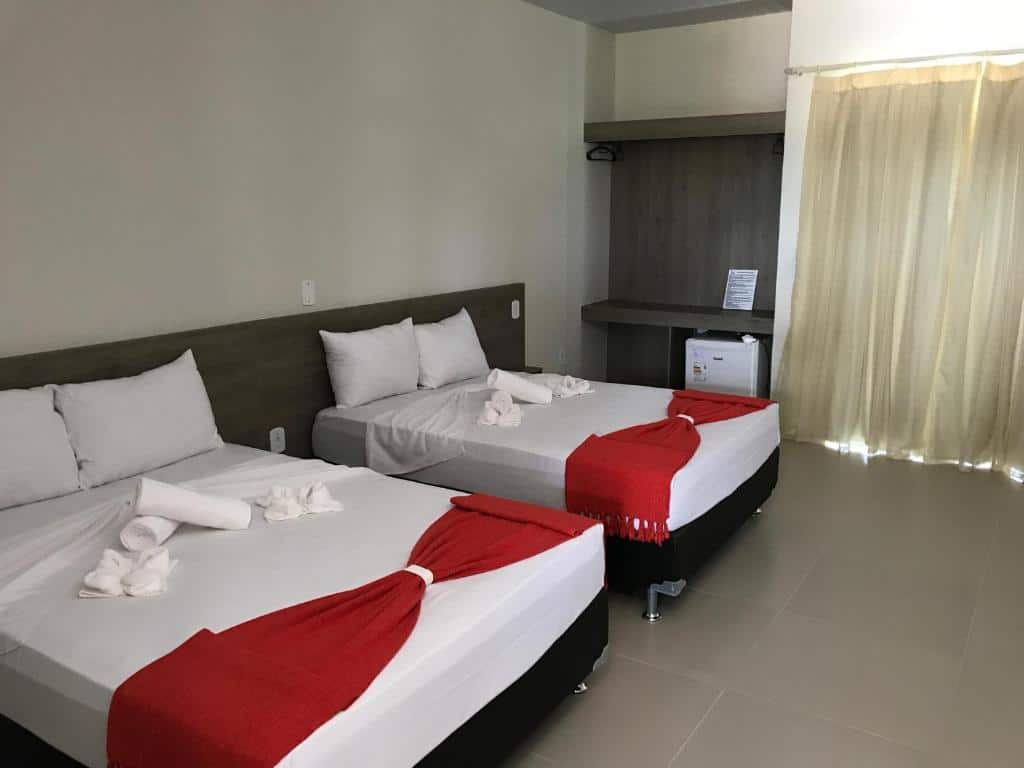 Duas camas de casal com roupa de cama branca e mantas vermelhas em um quarto, com escrivaninha, frigobar e cortinas bege, na Portal do Sol Lençóis Pousada, para representar pousadas em Barreirinhas.