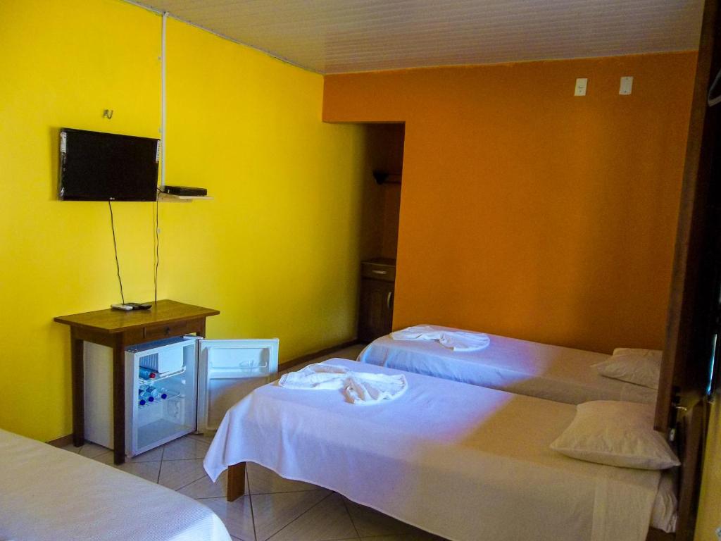 Um quarto na Toca do Vaidoso com três camas de solteiro, uma TV na parede, uma mesa pequena e um frigobar aberto embaixo da mesa, para representar pousadas em Barreirinhas.