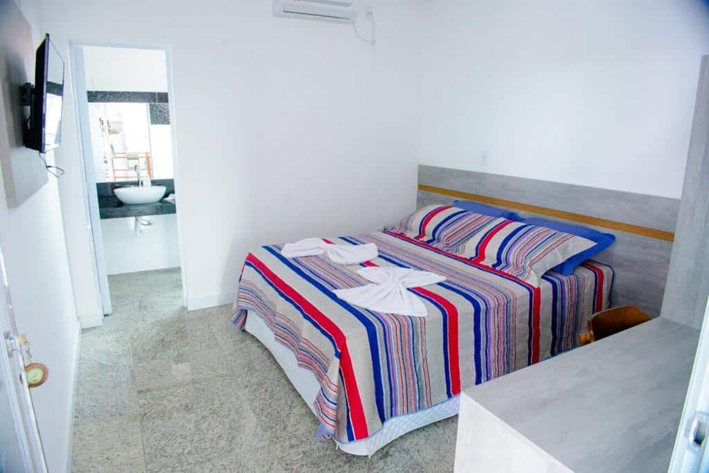 Um quarto simples com uma cama de casal, roupa de cama listrada, uma TV na parede e vista para um banheiro com pia, na Pousada Barreirinhas, para representar pousadas em Barreirinhas.