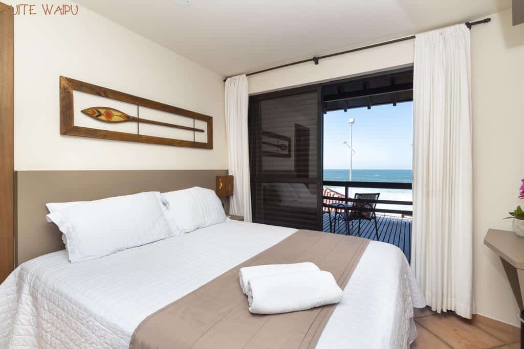 Interior do quarto da Pousada Kia Ora Bombinhas, de 10 m², com uma cama de casal com travesseiros e toalhas em cima, e uma varanda com chão de madeira, uma mesa redonda com duas cadeiras e vista para o mar azul e céu ensolarado