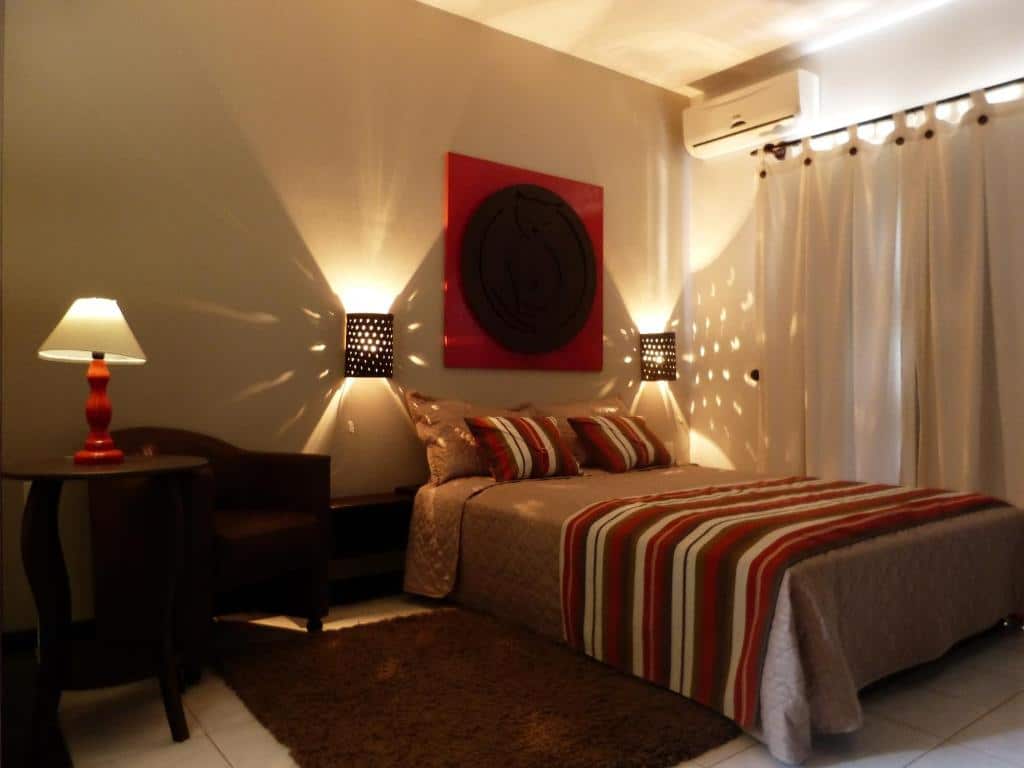 Interior do Quarto Deluxe da Pousada Narinari, com as cortinas fechadas e duas iluminarias (que ficam uma em cada lado da cama) iluminando o local com uma luz amarelada. A cama é de casal e em cima dela há dois travesseiros grandes, dois menores e um jogo de cama nas cores vermelho, marrom e branco. Ao lado há também uma poltrona marrom, uma mesa redonda com um abajur aceso e um ar-condicionado