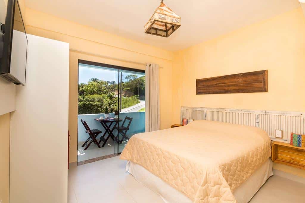 Suite, de 20 m², com cama queen-size da Vila Artesania. Além da cama, há também uma TV suspensa na parede, uma escrivaninha artesanal de cada lado da cama feita de caixotes e uma varanda com mesa quadrada e duas cadeiras com vista para a rua 