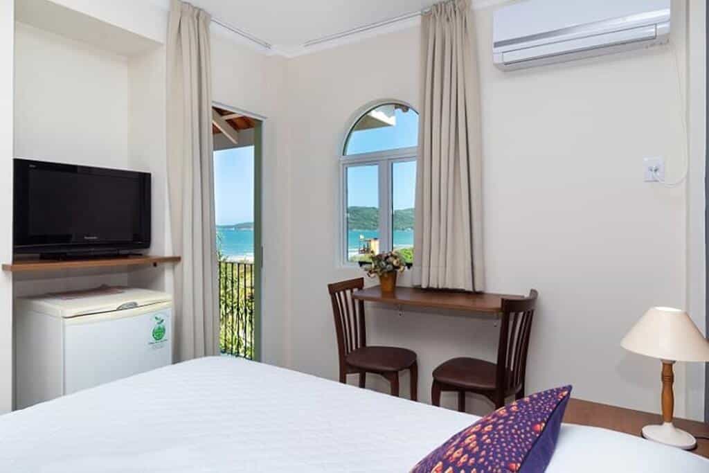Interior do Quarto Duplo da Pousada Villa dos Açores, de 18 m², com uma cama de casal, uma escrivaninha de madeira marrom escuro com um abajur branco ao lado, uma balcão pequeno com duas cadeiras, frigobar, TV e ar-condicionado para ilustrar as pousadas em Bombinhas. No lado direito, há uma porta dando acesso à varanda do quarto, que tem vista para o mar e montanhas da região