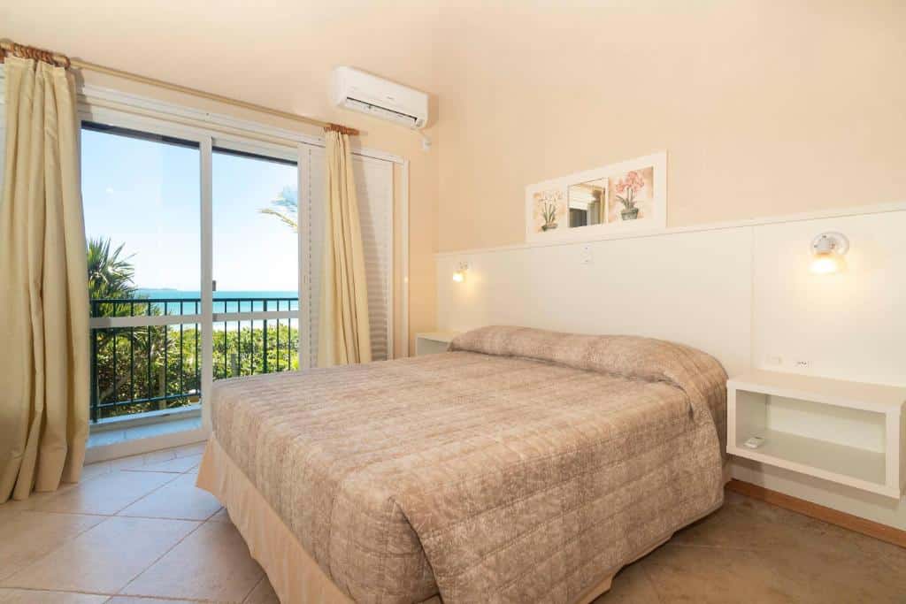 Interior do quarto da Pousada Morada do Guaruça, com uma cama de casal, ar-condicionado e sacada com vista para o mar azul e natureza verde localizada na orla da praia