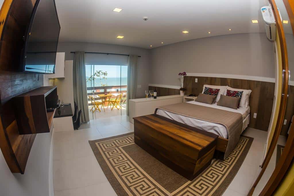 Interior da suíte da Pousada do Arvoredo, em Bombinhas, de 30 m², com uma cama de casal, TV e uma porta de vidro dando acesso à varanda do quarto, que tem uma mesa amarela com duas cadeiras e vista para o mar