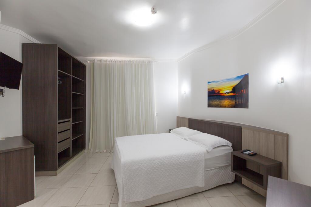 Interior do Quarto Duplo da Pousada Oásis de Mariscal, de 20 m², com as paredes brancas, um quadro com desenho de paisagem e pôr do sol, uma cama de casal, guarda-roupa e TV