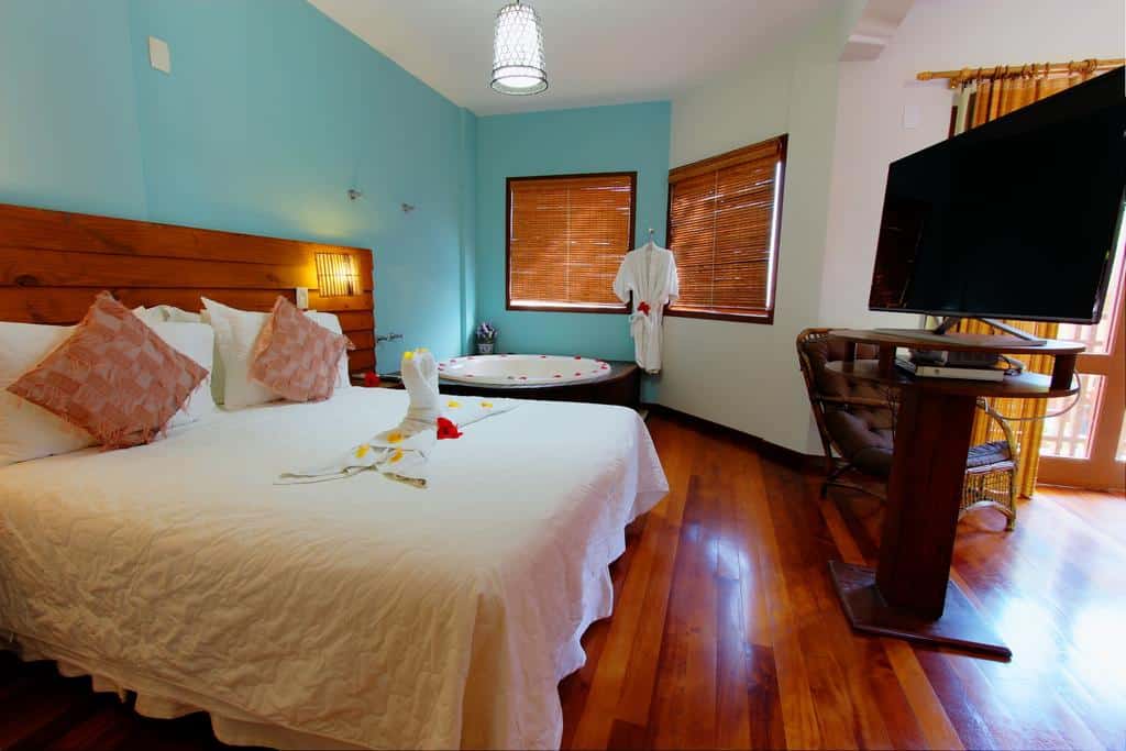 Interior de um dos quartos da Pousada Vila Boa Vida, com chão de madeira brilhando, cama de casal com escultura feita de toalha de banho e flores em cima, uma TV e, no lado direito, tem uma hidro redonda com pétalas vermelhas em volta e um roupão branco pendurado entre duas janelas