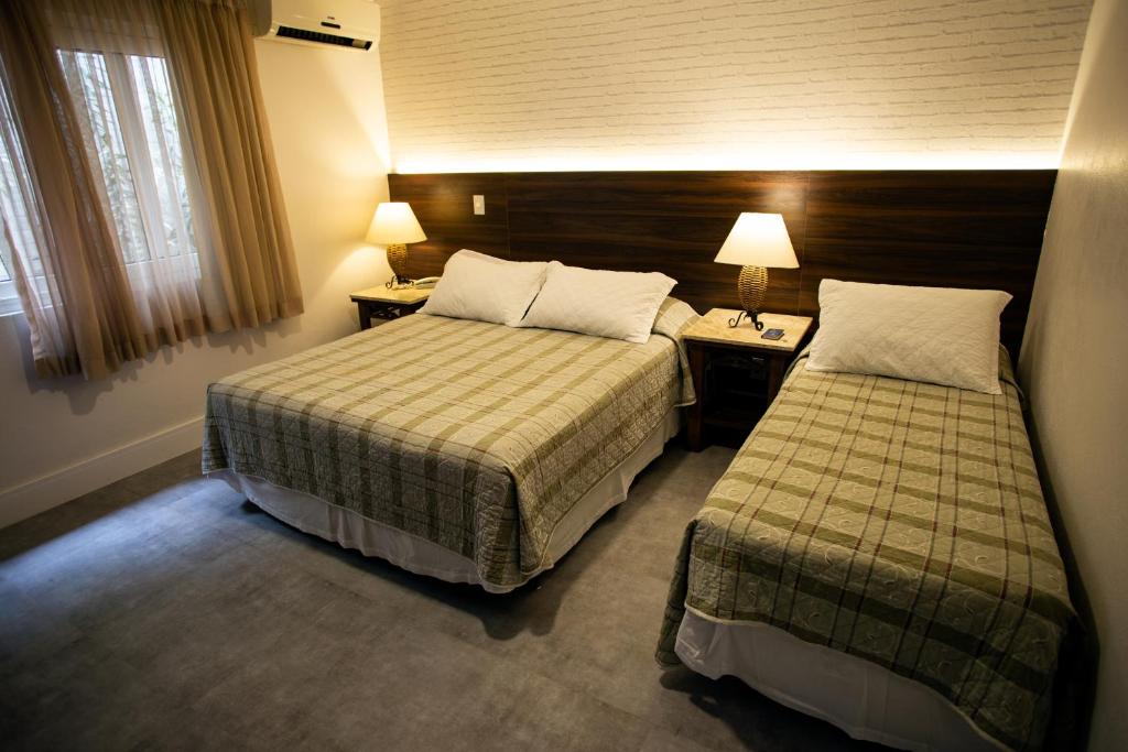 Um quarto no Pousada Águas Claras na Rua das Pedras, com uma cama de casal e uma cama de solteiro, duas mesas de cabeceira com luminárias e uma janela com cortinas bege, para representar pousadas em Búzios.