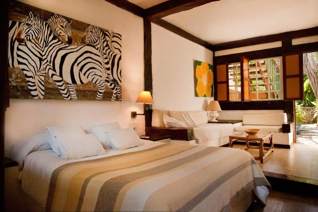 Um quarto com colcha listrada na Pousada Vila do Mar na Praia do Canto, pintura de zebra, sofás brancos, uma mesa de centro e janelas abertas com vista para o jardim, para representar pousadas em Búzios.