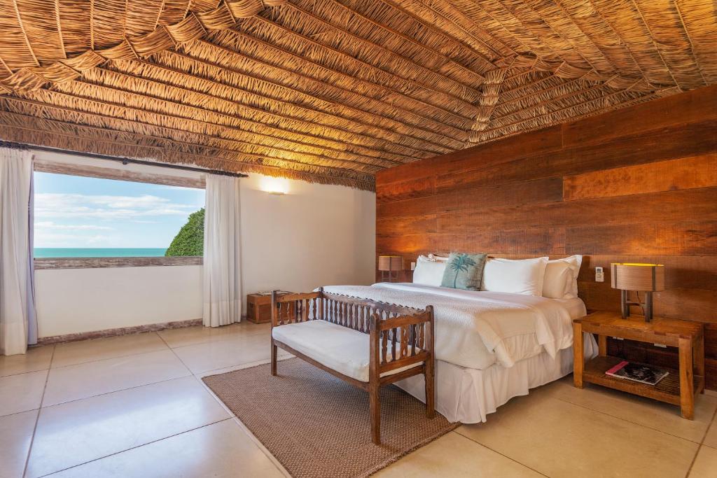 Um quarto na Pousada Villa Rasa na Praia Rasa com uma cama grande, banco de madeira, duas mesinhas de cabeceira, teto de tecido e uma janela com vista para o oceano, para representar pousadas em Búzios.