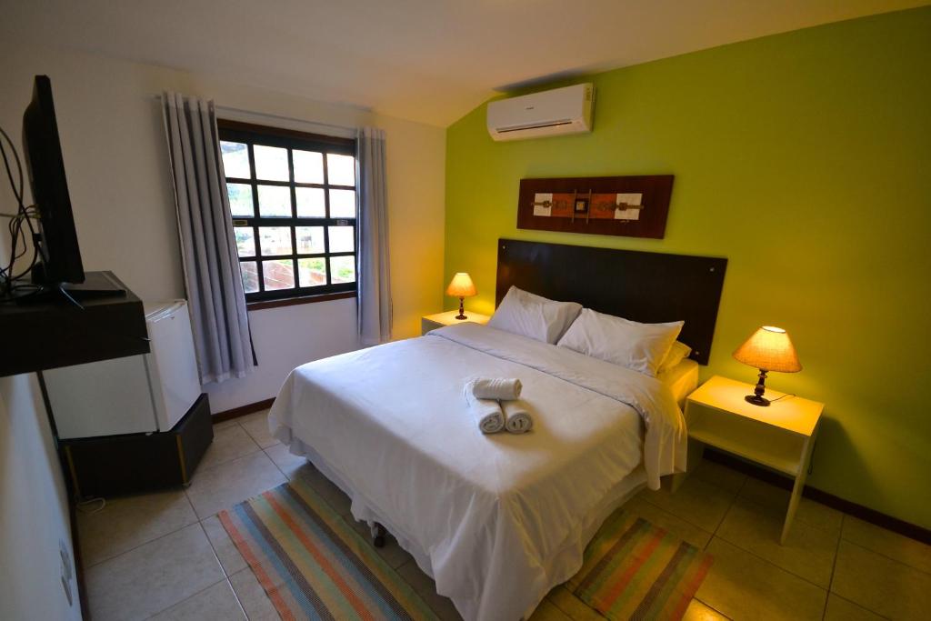 Uma cama bem arrumada com roupas de cama brancas, duas mesas de cabeceira com luminárias e uma parede verde de destaque em um quarto claro, em um dos quartos da Pousada do Centro, para representar pousadas em Búzios.