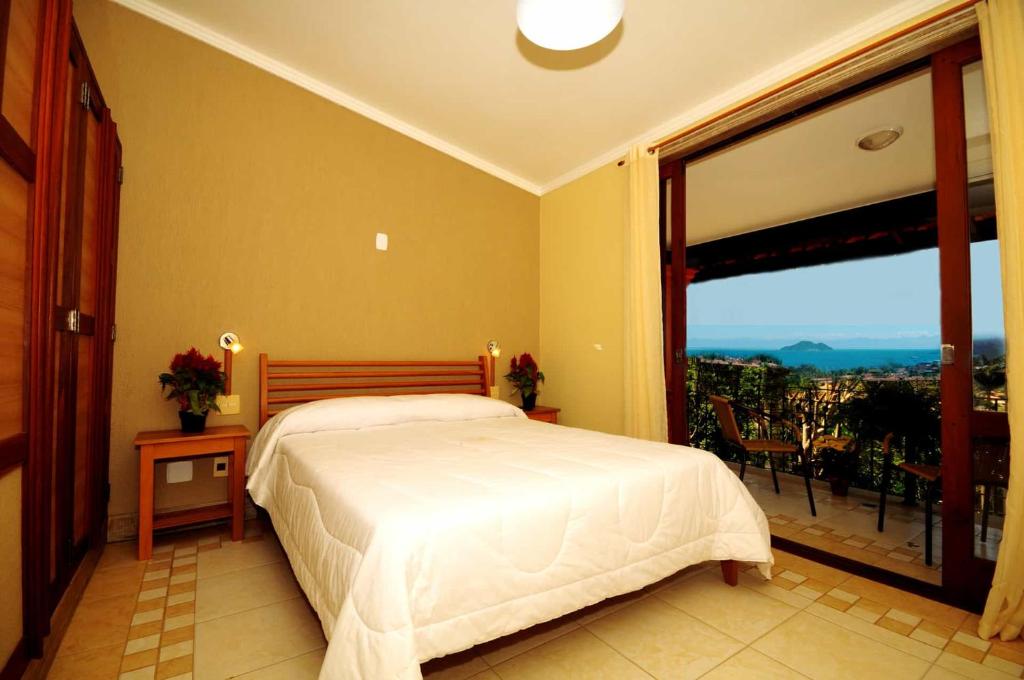 Um quarto com uma cama de casal, duas mesas de apoio e uma porta de vidro que se abre para uma varanda com vista para o mar, no quarto da Pousada Pelicano na Praia da Armação.