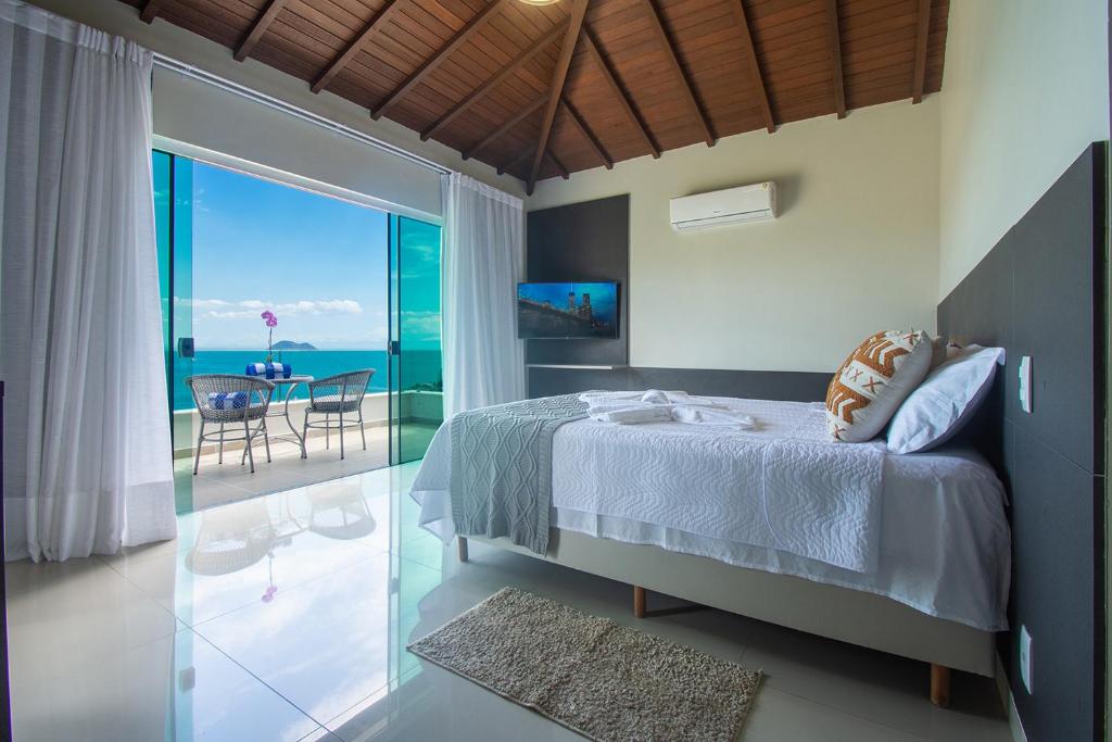 Um quarto moderno na Pousada Praia João Fernandes, com uma cama de casal, TV e portas de vidro que se abrem para uma varanda com vista para o mar e duas cadeiras, para representar pousadas em Búzios.