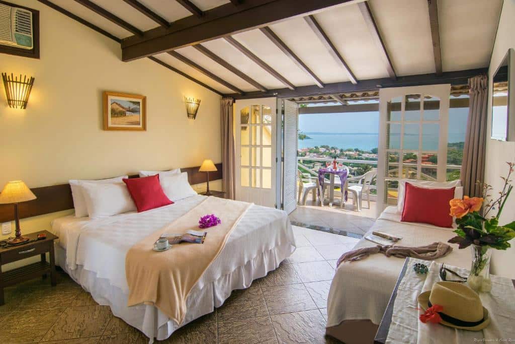 Quarto na Pousada e Spa Villa Mercedes na Praia João Fernandes, com uma cama de casal, cama de solteiro, flores e portas abertas que dão para uma varanda com vista para o mar, para representar pousadas em Búzios.