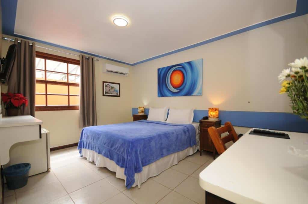 Uma cama bem arrumada com um cobertor azul, escrivaninha, cadeira e luminárias em um quarto bem iluminado e arrumado com uma janela, na Pousada El Parador no Centro, para representar pousadas em Búzios.