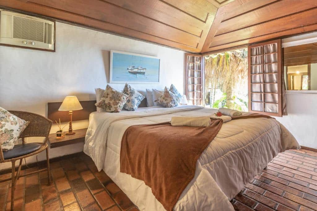 Um quarto aconchegante na Pousada Lestada na Praia dos Ossos, com uma cama de casal, cobertor marrom, travesseiros, abajur, cadeira, piso de tijolos e janela aberta com persianas de madeira, para representar pousadas em Búzios.