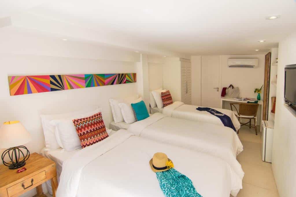 Um quarto na La Chimère Buzios Essence na Praia dos Ossos, bem iluminado com três camas de solteiro, travesseiros coloridos, uma escrivaninha, uma TV na parede e um chapéu em uma cama, para representar pousadas em Búzios.