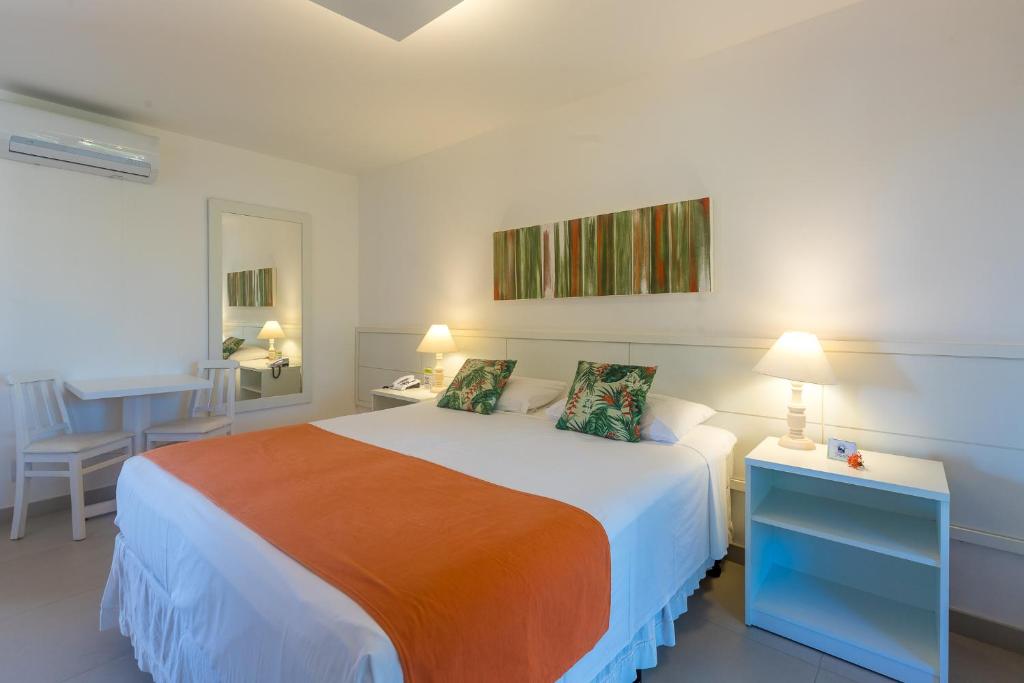 Um quarto da Pousada dos Tangarás na Praia de Geribá, com uma cama de casal, cobertor laranja, duas luminárias, uma pequena mesa com duas cadeiras e arte na parede acima da cama, para representar pousadas em Búzios.