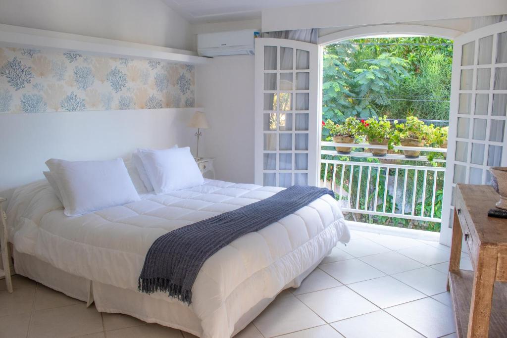 Uma cama bem arrumada em um quarto bem iluminado no Auberge de La Langouste na Praia da Armação com portas francesas abertas que levam a uma varanda com vasos de plantas e vegetação do lado de fora, para representar pousadas em Búzios.