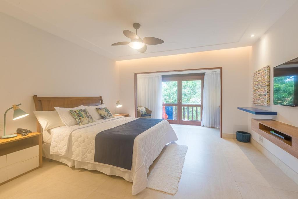 Um quarto bem iluminado no Corais e Conchas na Praia de Geribá, com uma cama grande, duas mesas de cabeceira, um ventilador de teto, TV e portas de vidro que levam a uma varanda, para representar pousadas em Búzios.
