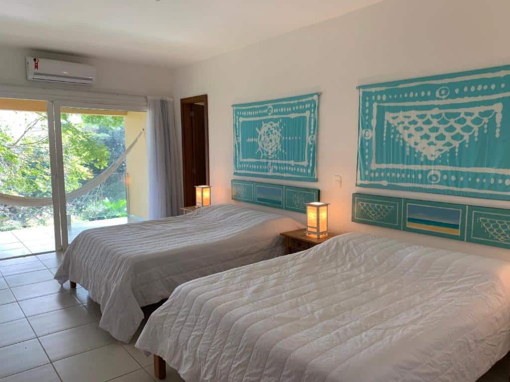 Um quarto bem iluminado na Praia da Ferradurinha Guest House, com duas camas, roupa de cama branca, arte na parede azul, luminárias de cabeceira e uma rede do lado de fora de uma porta de vidro deslizante, para representar pousadas em Búzios.