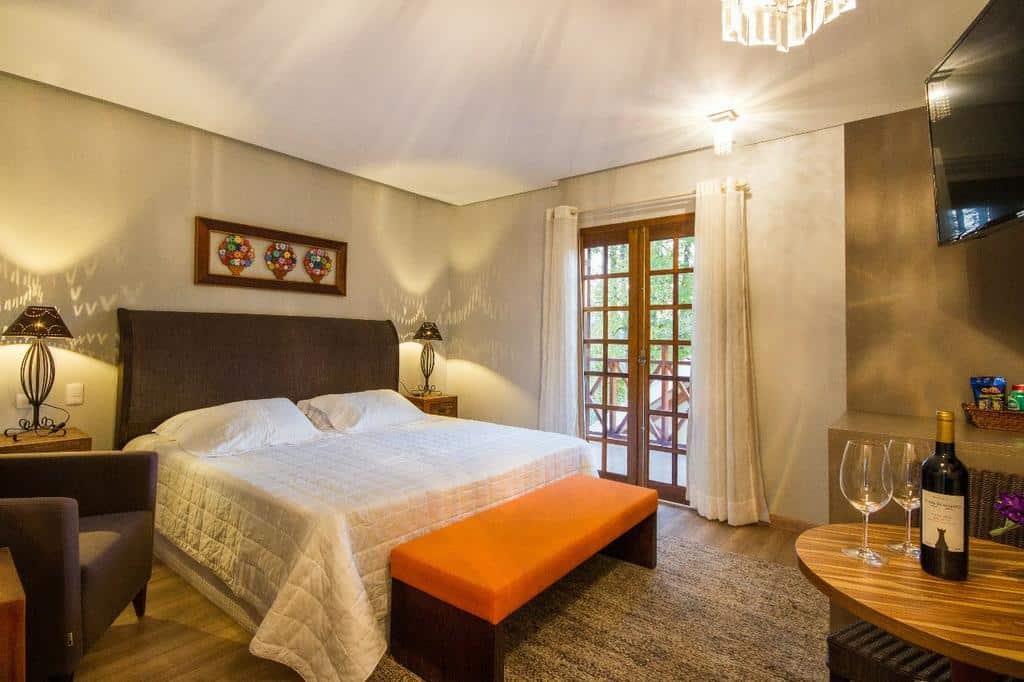 Quarto na Pousada Cantinho da Serra bem iluminado com cama king size, banco laranja, portas de sacada e uma mesa posta com vinho e taças, para representar pousadas em Campos do Jordão.