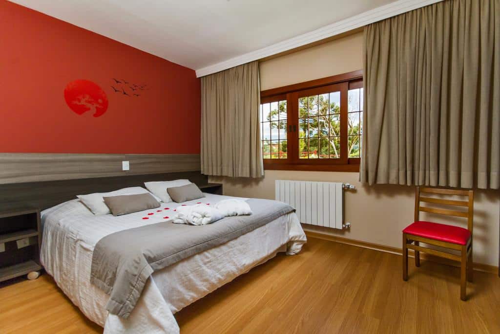 Uma cama bem arrumada com roupa de cama cinza e branca em um quarto com parede com detalhes em vermelho, janela e cadeira de madeira, na Matsubara Pousada, para representar pousadas em Campos do Jordão.