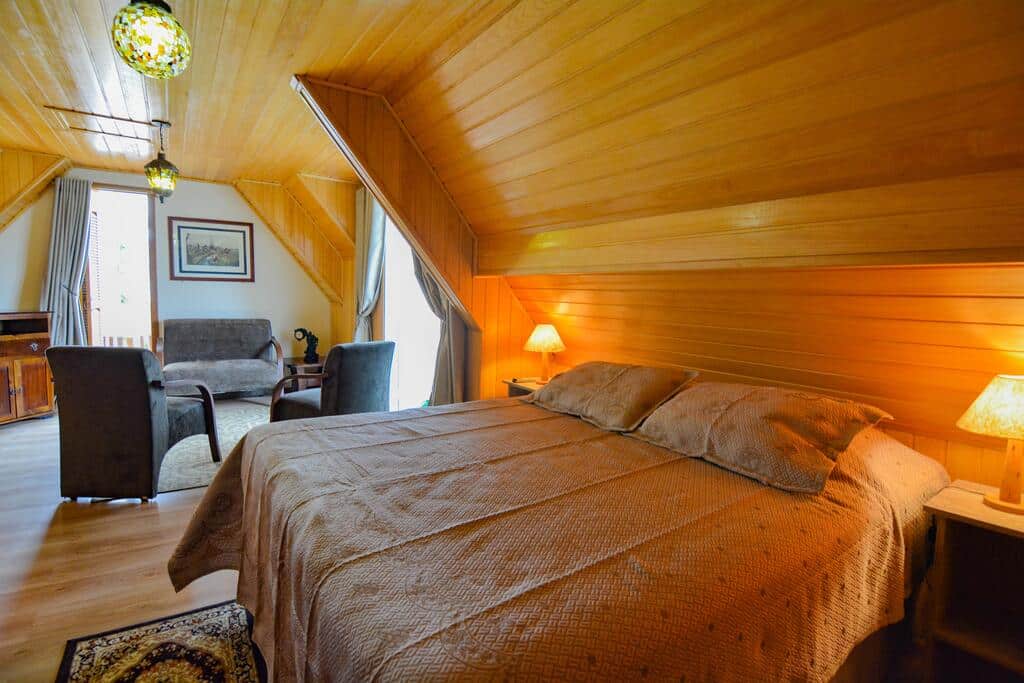 Um quarto aconchegante na Pousada Kaliman, com paredes com painéis de madeira, uma cama de casal, duas luminárias, cadeiras e uma pequena área de estar, para representar pousadas em Campos do Jordão.